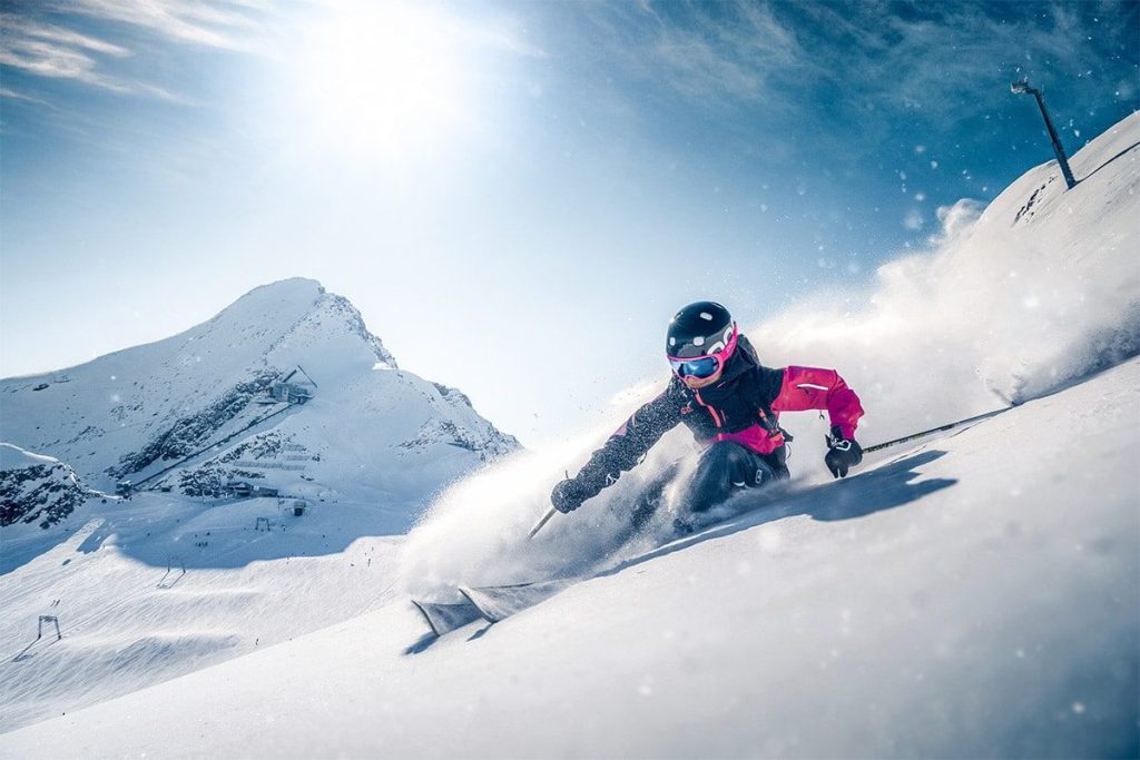 Skifahrer auf verschneiter Piste mit Bergkulisse und strahlendem Himmel im Hintergrund.