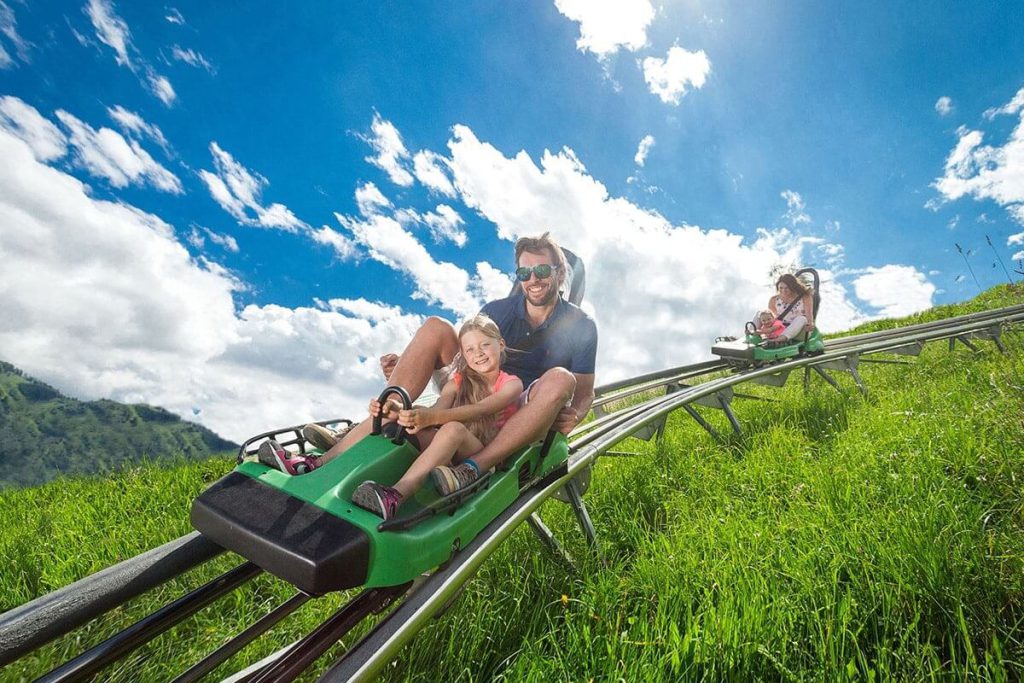 Familie fährt lachend mit Sommerrodelbahn durch die grüne Landschaft bei sonnigem Wetter.