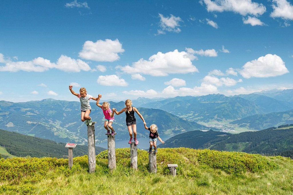 Kinder springen fröhlich auf Baumstämmen mit Bergblick in Zell am See.