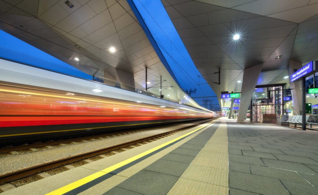 Moderne Bahnhofshalle bei Nacht, Zug fährt schnell vorbei, beleuchtete Plattform und Architektur.