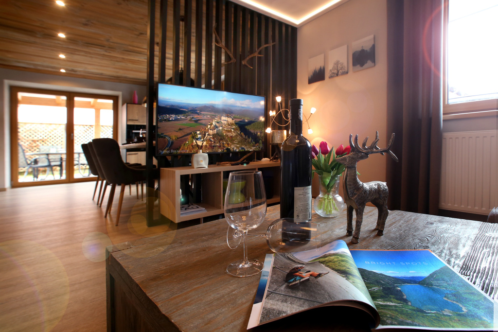 Część dzienna apartamentu z drewnianym stołem, kieliszkami do wina, butelką, telewizorem, jadalnią i drzwiami tarasowymi